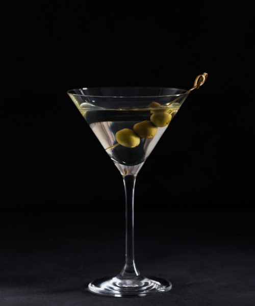 Glass of classic dry martini cocktail with olives on dark stone table against black background.